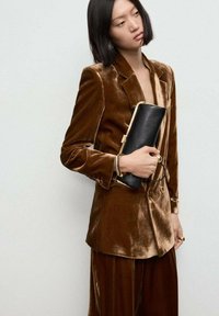 Bruine fluwelen blazer met een notch-lapel, voorzien van een zak; gecombineerd met bijpassende hoge taillebroeken. Model houdt een zwarte clutch met gouden accenten vast.