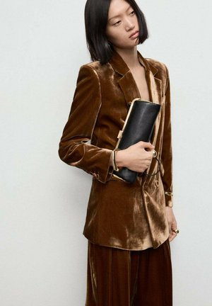 Blazer de terciopelo marrón con solapa a rayas, que cuenta con un bolsillo; combinado con pantalones de cintura alta a juego. La modelo sostiene un bolso negro con detalles dorados.