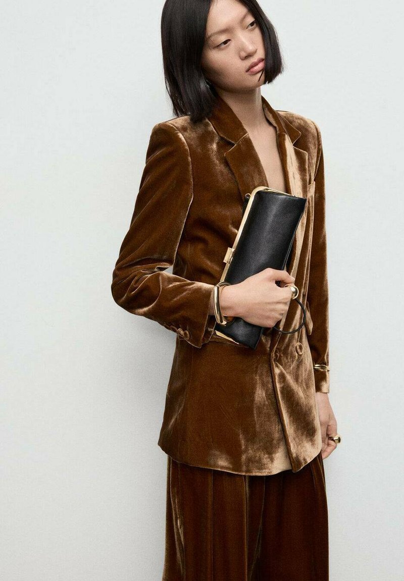 Bruine fluwelen blazer met een notch-lapel, voorzien van een zak; gecombineerd met bijpassende hoge taillebroeken. Model houdt een zwarte clutch met gouden accenten vast.