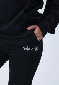 Sudadera y pantalones negros en un material suave, con bordado blanco "Project x Paris" en los pantalones. Diseño ajustado con puños acanalados.