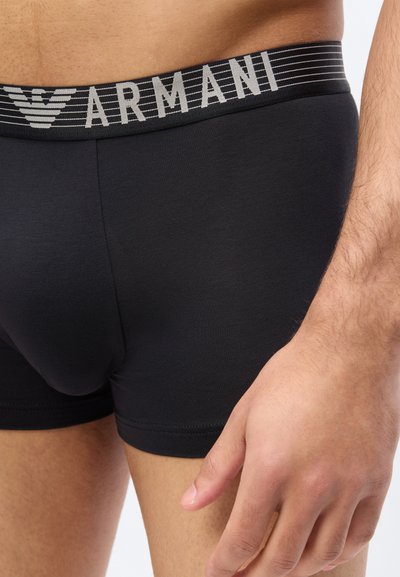 Ανδρικός κορμός και χέρι που φορούν μαύρα ανδρικά μποξεράκια Armani με γκρι ριγέ μέση και λογότυπο σε λευκό φόντο.