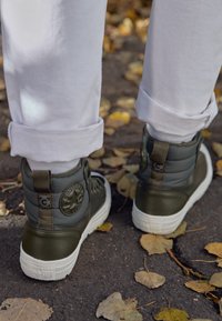 Zapatillas altas de color verde oliva con parte superior de tela texturizada, suela de goma blanca y parche de logo icónico en el talón. Pantalones blancos enrollados cerca.