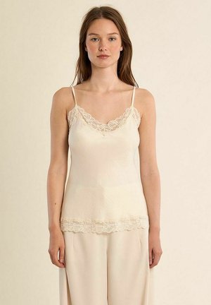 Giovane donna che indossa un top crema con pizzo e pantaloni larghi color crema, in piedi davanti a uno sfondo beige chiaro uniforme.