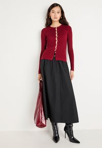 Raudona ilgomis rankovėmis megztinis su sagų uždarymu, derinamas su juoda maksi sijonu ir juodais aštriakampiais pasmaugtiniais batais. Bordo spalvos rankinė.