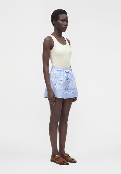 Femme debout portant un haut beige sans manches, un short bleu clair brodé avec un cordon et des sandales marron à boucle sur un fond uni.