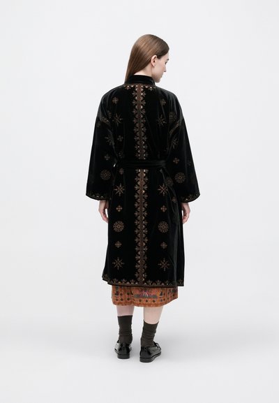 Juodas aksominis chalatas su auksinių siuvinėjimų raštais, ilgomis rankovėmis ir liemens diržu. Modelis dėvi po juo rudą raštuotą sijoną.