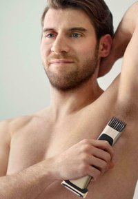 Tondeuse électrique avec un corps métallique, une prise en caoutchouc noir et un peigne. Un homme l'utilise en levant son bras, révélant des poils sous les bras.