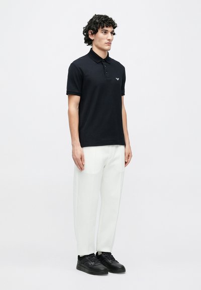 Čierne polo tričko s golierom, krátkymi rukávmi a malým logom na hrudi, v kombinácii s bielymi voľnými nohavicami a čiernymi teniskami.