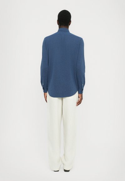 Chemise bleue à manches longues en tissu texturé avec un ourlet incurvé, assortie à un pantalon large blanc. Détails de boutons aux poignets.
