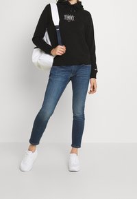 Osoba oblečená v černé mikině Tommy Jeans, modrých skinny džínách, bílých teniskách a nese bílý batoh na jednom rameni.