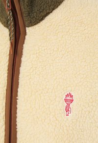 Fleecejacke in Creme mit olivfarbenem Kragen, ausgestattet mit einem braunen Reißverschluss und einem roten, gestickten Fackel-Logo auf der linken Seite. Texturiert.