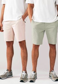 Deux hommes portant des t-shirts blancs, des shorts pastel rose et vert, et des baskets grises New Balance, debouts les mains dans les poches sur un fond blanc.