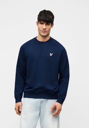 Moški, oblečen v temno modro jopico z majhnim belim logotipom ptice na prsih in svetlo moderimi jeans hlačami, stoji pred enobarvnim ozadjem.