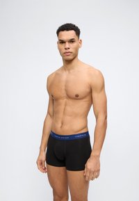 Άνδρας χωρίς πουκάμισο όρθιος, φορώντας μαύρα μπόξερ με μπλε μέση με το λογότυπο "Tommy Hilfiger" σε απλό γκρι φόντο.