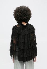 Sort tylkappe med lagdelte flæser, en høj flæset krave og gennemsigtigt stof. Modellen har grå metallicbukser på.