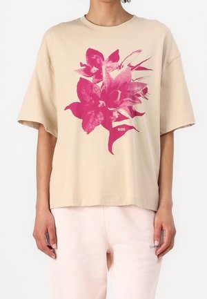 Beige oversized t-shirt met grote roze bloemenprint en "BOSS" tekst, gedragen met lichtroze broek.