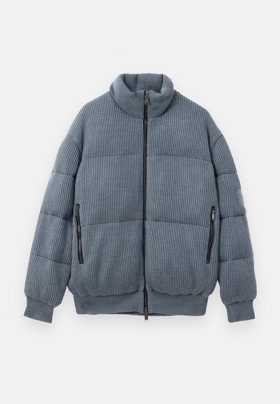 Chaqueta gris acolchada con textura estriada, cuello alto y bolsillos con cremallera negros. Presenta una cremallera frontal completa y puños elásticos.