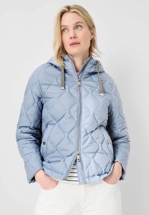 Frau trägt hellblaue gesteppte Kapuzenjacke mit schwarz-weißen Kordeln, Hände in den Taschen, schaut leicht nach rechts vor einfarbigem Hintergrund.