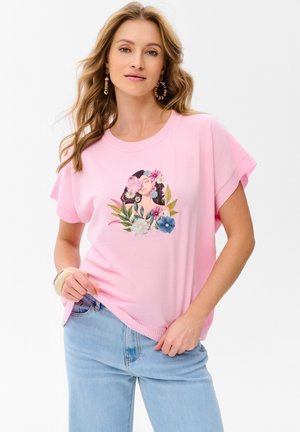 Kobieta o falowanych włosach, ubrana w różową koszulę z kwiatową ilustracją kobiety oraz jasnobłękitne dżinsy, pozująca na białym tle.