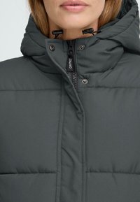 Tamno siva puffer jakna s visokim ovratnikom, patentnim zatvaračem i kopčama na dugmad, podesivom vezicom na kapuljači i quilted teksturom.