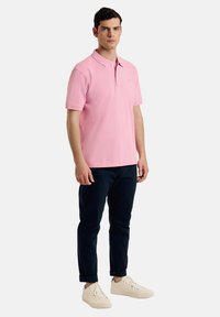Polo de algodón rosa con cuello clásico, mangas cortas y un pequeño logo bordado, combinado con chinos azul marino y zapatillas blancas.