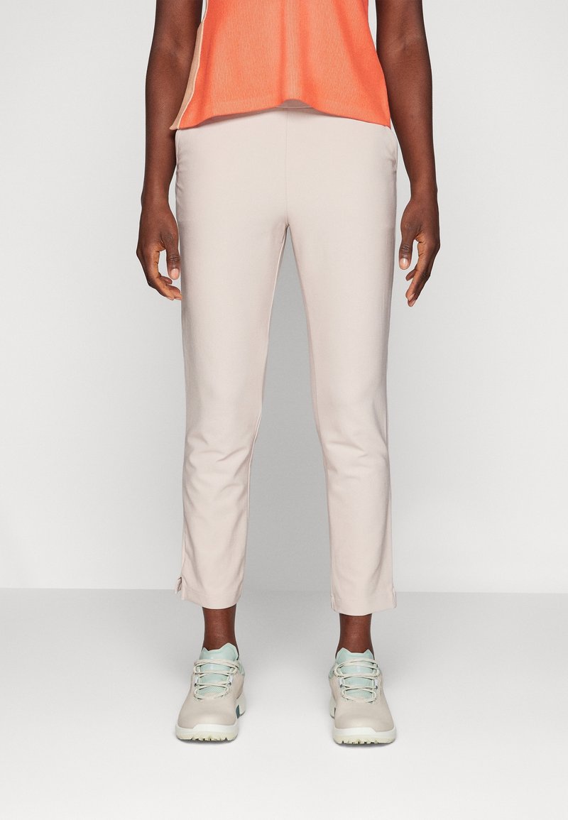 Personne portant un pantalon beige slim, des baskets bleu clair et beige, et un haut sans manches orange, debout devant un fond uni.