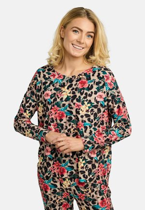 Nasmejana žena s plavom kosom dužine do ramena, nosi uparenu cvjetnu i leopard uzorku bluzu s dugim rukavima i hlače, ruke opušteno sklupčane.