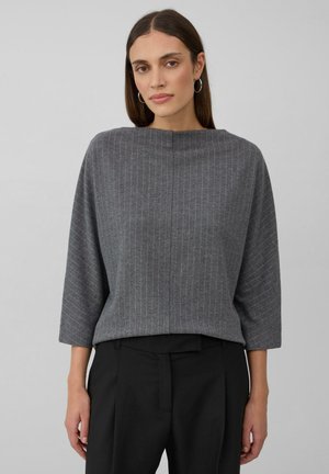Vrouw gekleed in een grijs streepdessins bootnecktop met driekwart mouwen en zwarte hoge taille broek, staand tegen een plain achtergrond.