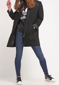 Svart kappa med skräddarsydd design, öppen front och långa ärmar, bärs över en mörk sweatshirt och blå jeans, i kombination med svarta sneakers.