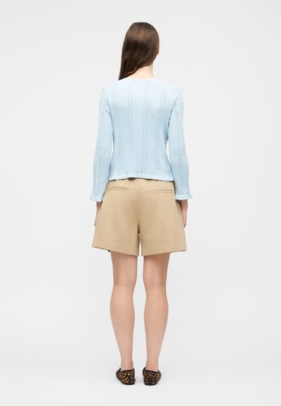 Γυναίκα με μακριά καστανά μαλλιά, φορώντας ανοιχτό μπλε μπλουζάκι με μακριά μανίκια, μπεζ σορτς μέχρι το γόνατο και μοκασίνια με λεοπάρ σχέδιο, στέκεται με την πλάτη προς τα εμπρός.