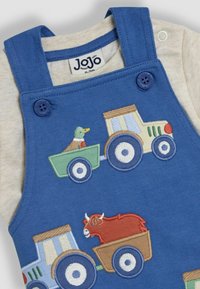 Salopette en coton bleu avec des tracteurs brodés, présente un camion vert avec un canard et un camion marron avec un taureau rouge. Boutons en plastique sur les bretelles.