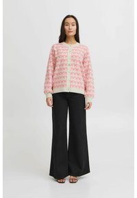 Gilet en tricot rose pâle avec des motifs de nœuds rouges répétés, présentant un devant à boutons et des poignets côtelés, associé à un pantalon noir évasé.