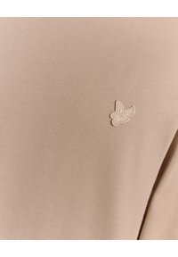 Beige pullover lavet af blødt stof med et lille, broderet fuglelogo i en lidt lysere nuance på fronten.
