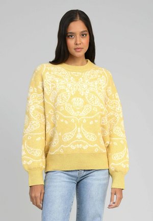 Jeune femme aux longs cheveux bruns portant un pull jaune avec des motifs cachemire blancs et un jean bleu clair, sur un fond gris uni.