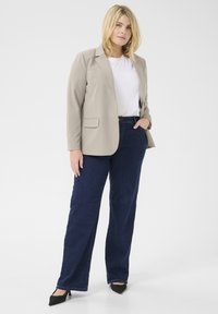 Blazer beige sopra una maglietta bianca, abbinato a jeans blu scuro. Il blazer ha una vestibilità sartoriale e due tasche frontali. Tacchi a spillo neri.