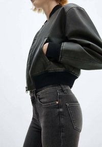 Chaqueta bomber verde oscuro con puños y dobladillo negro acanalados, con cierre de cremallera y bolsillos laterales, combinada con jeans gris oscuro.