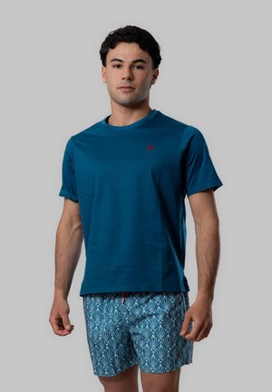 Giovane uomo con una maglietta blu tinta unita e pantaloncini blu con fantasia, in piedi davanti a uno sfondo grigio chiaro uniforme.