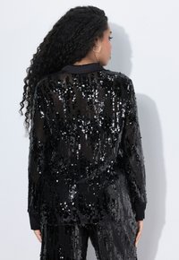 Black sequin blazer with vertical stripes and long sleeves, featuring a solid black collar and cuffs, creating a textured, shimmering surface.