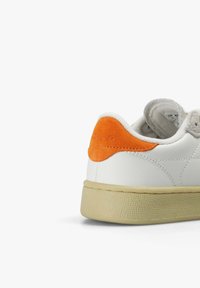 Baskets blanches avec semelle beige et languette de talon en daim orange, coutures visibles et col rembourré à la cheville sur un fond blanc.