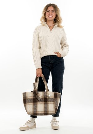 Kvinde smilende, iført creme kabelstrikket trøje, mørke jeans, hvide sneakers, holder stor brun og creme ternet tote taske med hank.