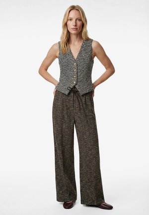 Blonde vrouw die een mouwloze grijze gebreide vest met knopen draagt, brede donkere broek met patroon en donkere schoenen, staand met handen achter de rug.