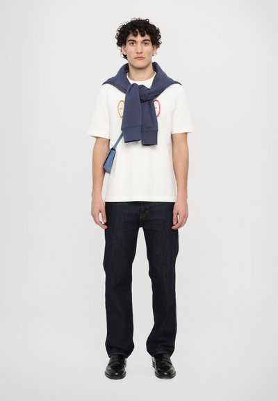 Bela majica s pisanimi okroglimi vezeninami, mornarsko modra jopica povezana okoli vratu, temno denim hlače in črne čevlje.