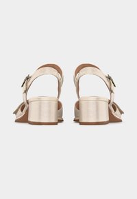 Ζευγάρι μπεζ ανοιχτά σανδάλια με χοντρά τακούνια και λουράκια στον αστράγαλο με πλαϊνές αγκράφες, από πίσω σε απλό φόντο.