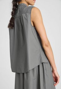 Blusa de tejido gris sin mangas con un alto collar, que presenta un detalle de espalda fruncido y un corte relajado, cayendo suavemente sobre los hombros.