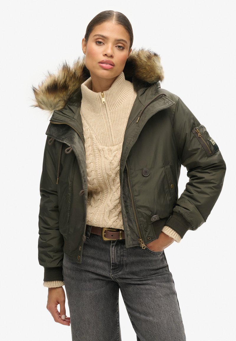 Olijfgroene pufferjack met een nepbont capuchon, twee voorkruiszakken en een ritssluiting aan de voorkant, gecombineerd met een crèmekleurige kabelgebreide trui.