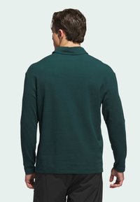 Dark green long-sleeve polo shirt with a collar, smooth texture, and a relaxed fit. Features side slits at the hem for ease of movement.