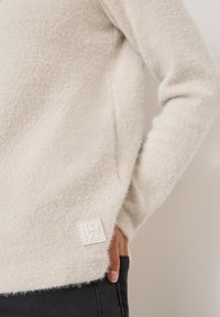 Kremowy sweter z dzianiny o teksturowanym wykończeniu, z małą białą metką z napisem "COZY" na dole. Miękki, luźny krój.