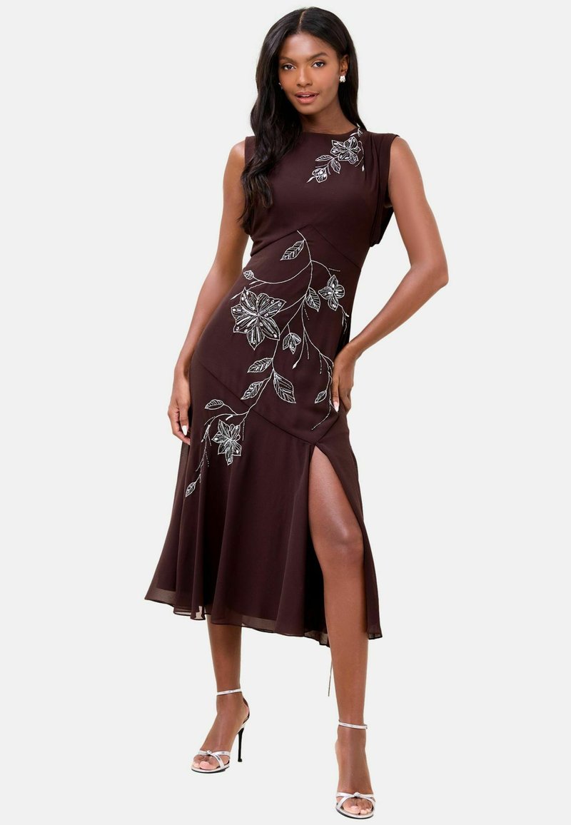 Femeie purtând o rochie midi maro închis, fără mâneci, cu broderie florală albă și o deschizătură pe coapsă, pozând cu mâna pe șold și purtând pantofi cu toc înalt.