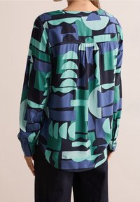 Chemise à manches longues aux motifs géométriques verts et bleus, avec une coupe décontractée, un ourlet arrondi et des poignets boutonnés. Texture de tissu lisse.