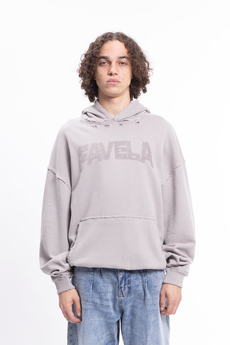 Giovane con capelli ricci indossa un hoodie grigio chiaro logorato con la scritta "FAVELA" e jeans blu larghi, in piedi davanti a uno sfondo bianco semplice.
